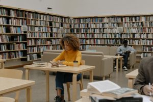 Students studying in a university library surrounded by bookshelves and tables.